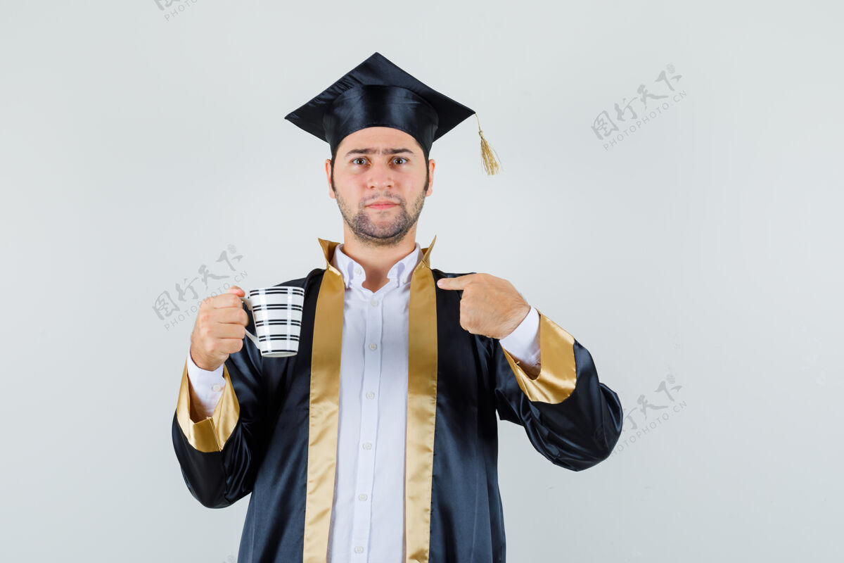 胡茬年轻人手里拿着一杯饮料 指着毕业制服前的自己站立毕业制服