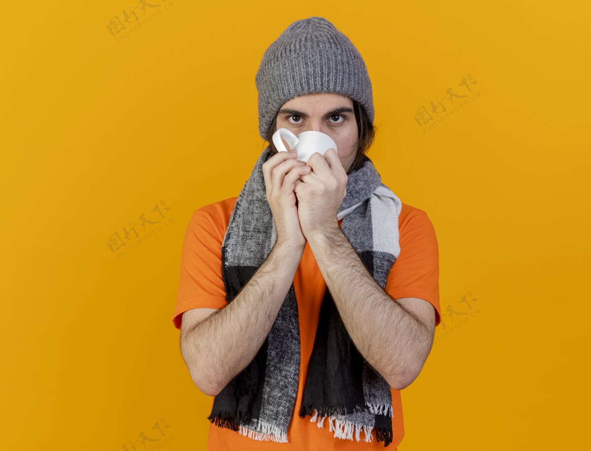 生病看着镜头年轻的病夫戴着冬天的帽子戴着围巾喝茶隔离在橙色的背景上帽子围巾年轻人