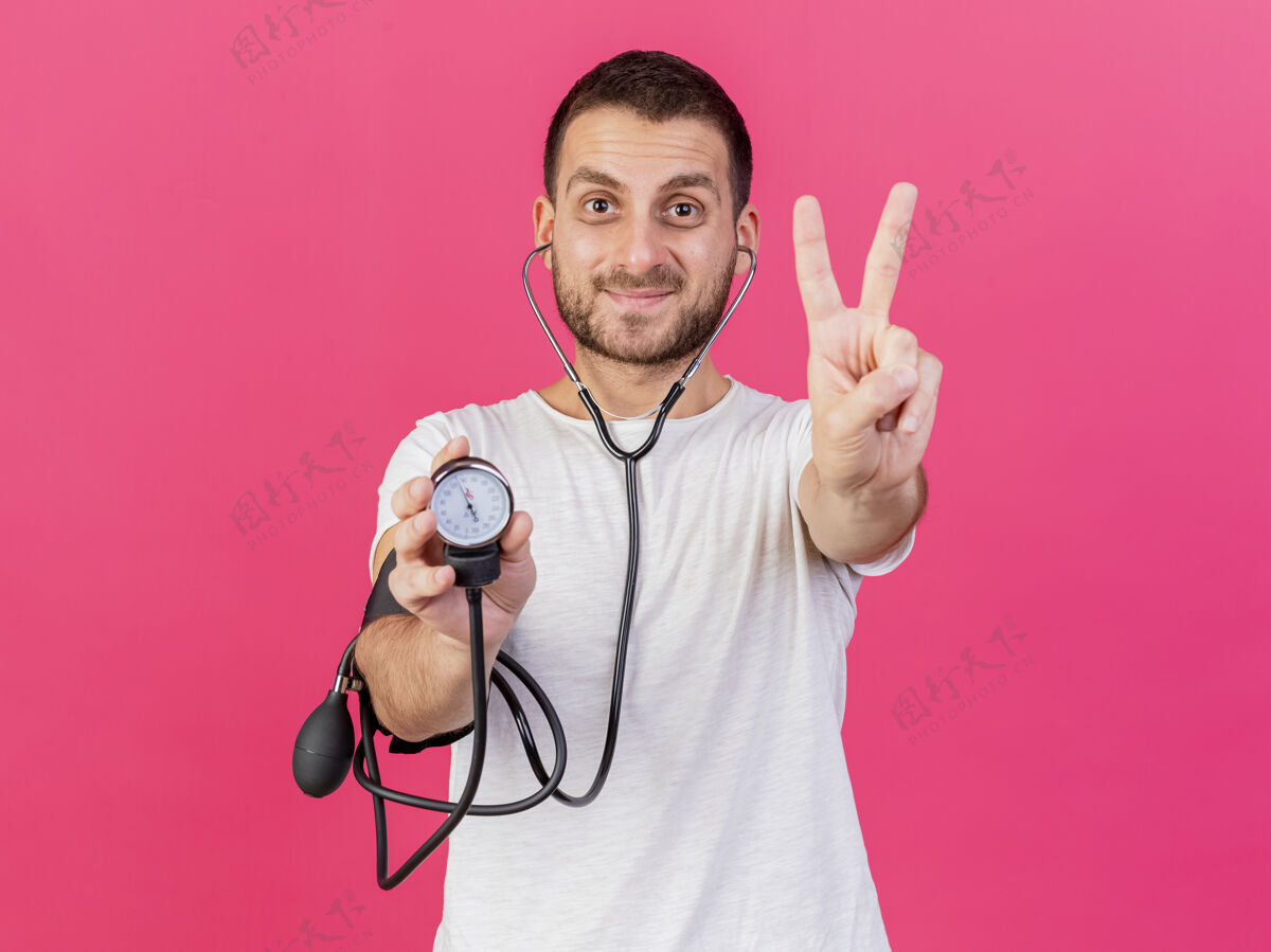 请戴着听诊器拿着血压计的年轻病患高兴地表示和平姿态隔离在粉红色背景上姿势听诊器年轻