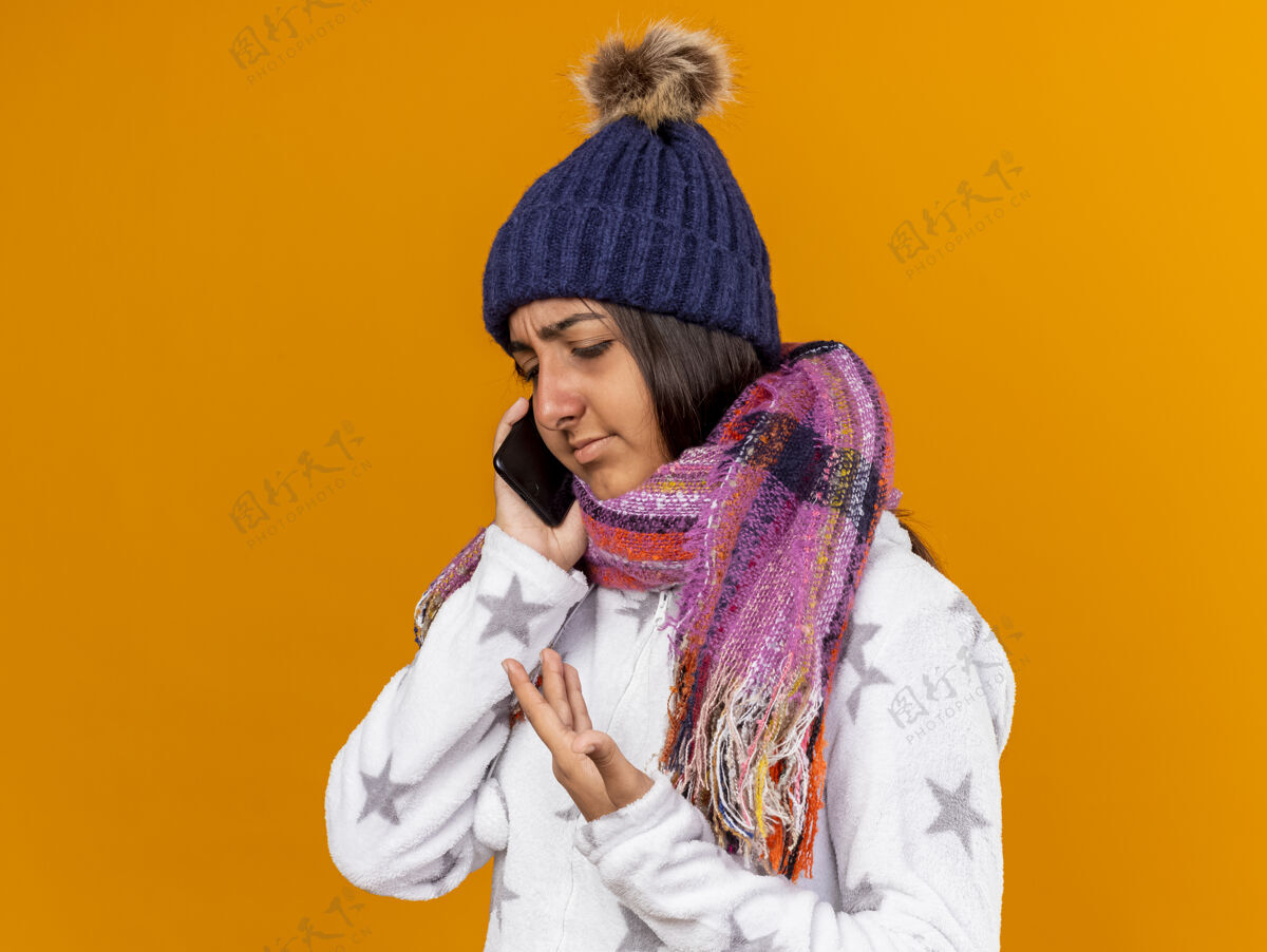不愉快戴着围巾的冬天帽子的未出狱的生病的年轻女孩在电话里讲话 黄色背景下被隔离说话疾病帽子