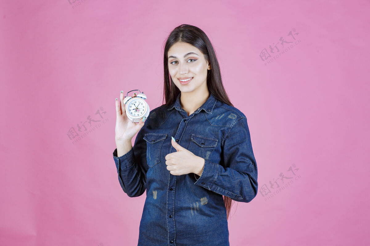 模特穿着牛仔衬衫的年轻女子拿着闹钟 做着好手势工人年轻积极