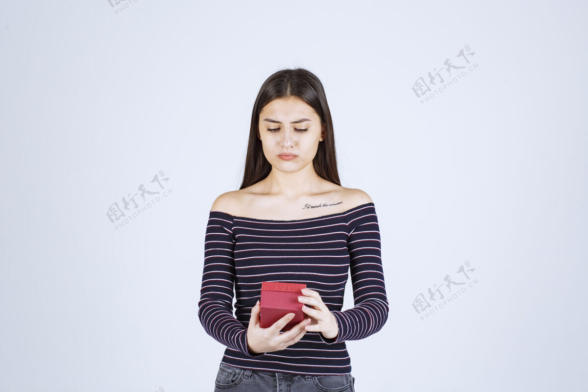 休闲穿着条纹衬衫的女孩手里拿着一个红色的礼盒 看上去既困惑又怀疑意外员工姿势