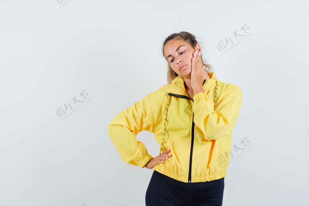 头发金发女人面颊靠在手掌上 手放在腰上 穿着黄色棒球服和黑色裤子 看起来很沉思金发美女裤子