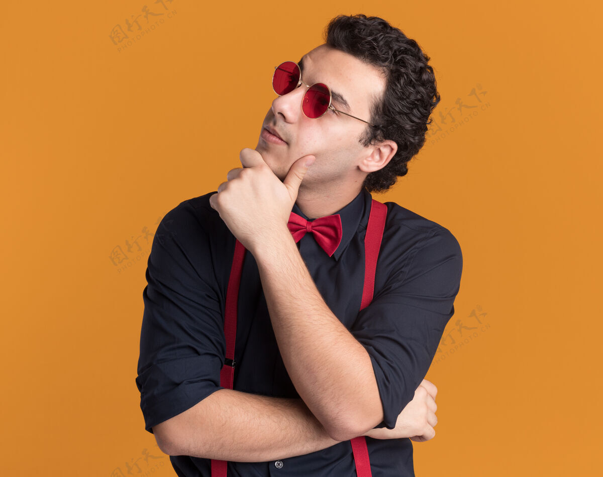穿戴着蝴蝶结 戴着眼镜 系着吊带 手放在下巴上 站在橙色墙壁上思考的时髦男人时尚思考靠边