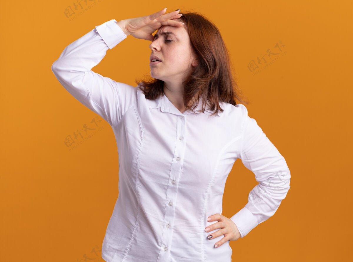 女人一个穿着白衬衫 手放在头上的年轻女人站在橘色的墙上又累又无聊年轻衬衫无聊