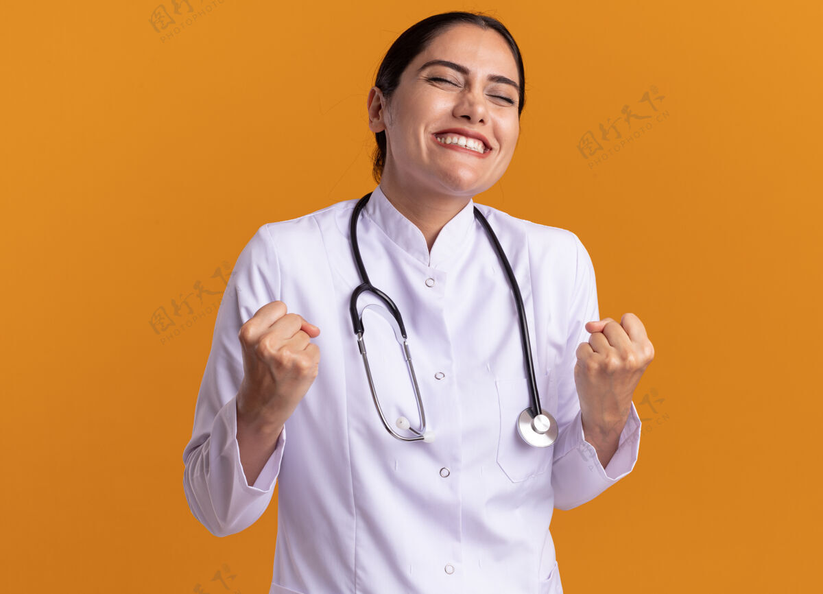 女人身穿医用外套 脖子上戴着听诊器 握紧拳头的年轻女医生站在橘色的墙上 既高兴又兴奋握紧拳头兴奋