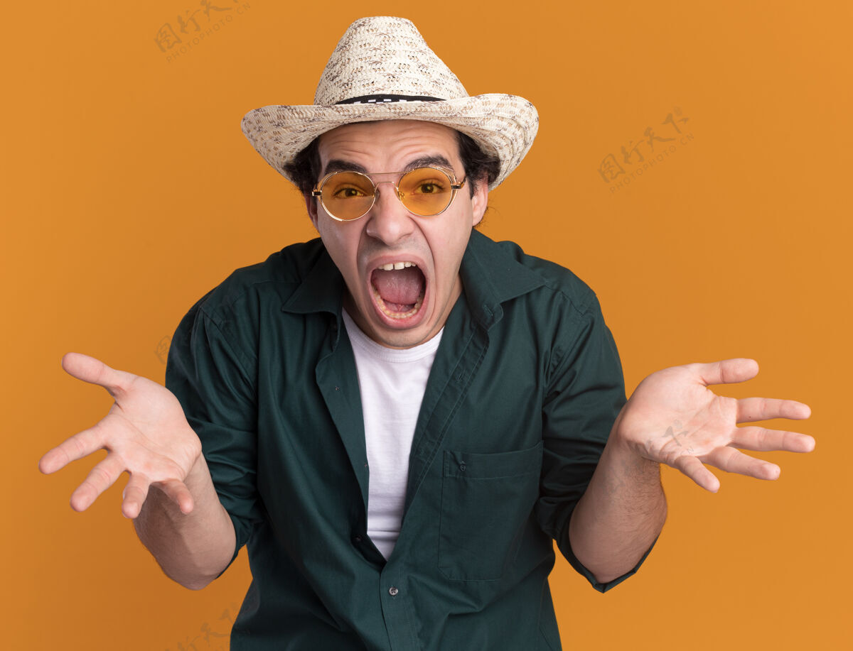 眼镜一个穿着绿衬衫戴着夏令帽戴着眼镜的年轻人站在橘色的墙上 看着前面狂吼着喊着男人举着