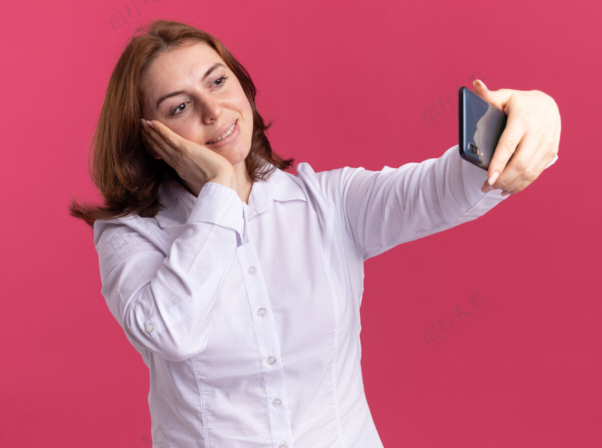年轻穿着白衬衫的快乐年轻女子用智能手机自拍 站在粉红色的墙上开心地微笑着女人欢呼手机