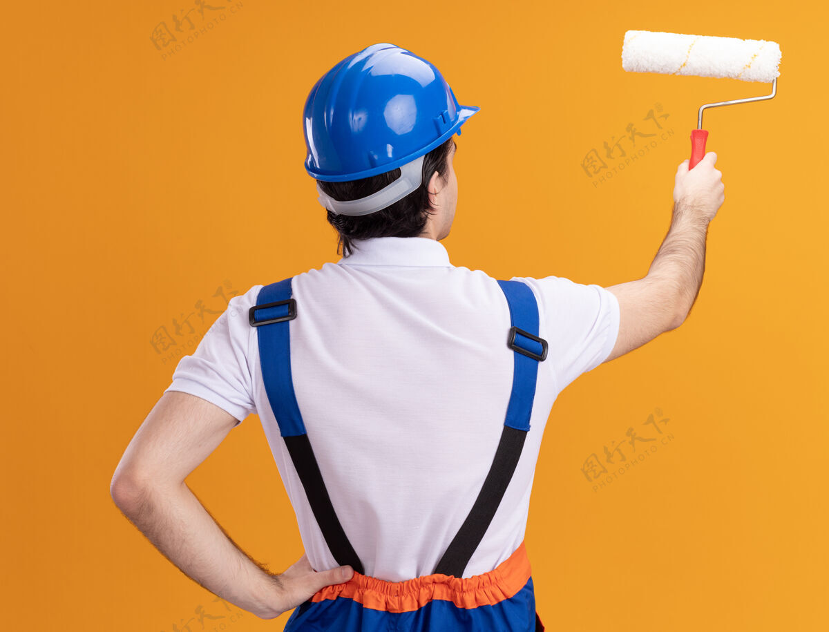 滚轴年轻的建筑工人穿着建筑制服 戴着安全帽 背着油漆辊站在橙色的墙上头盔背安全