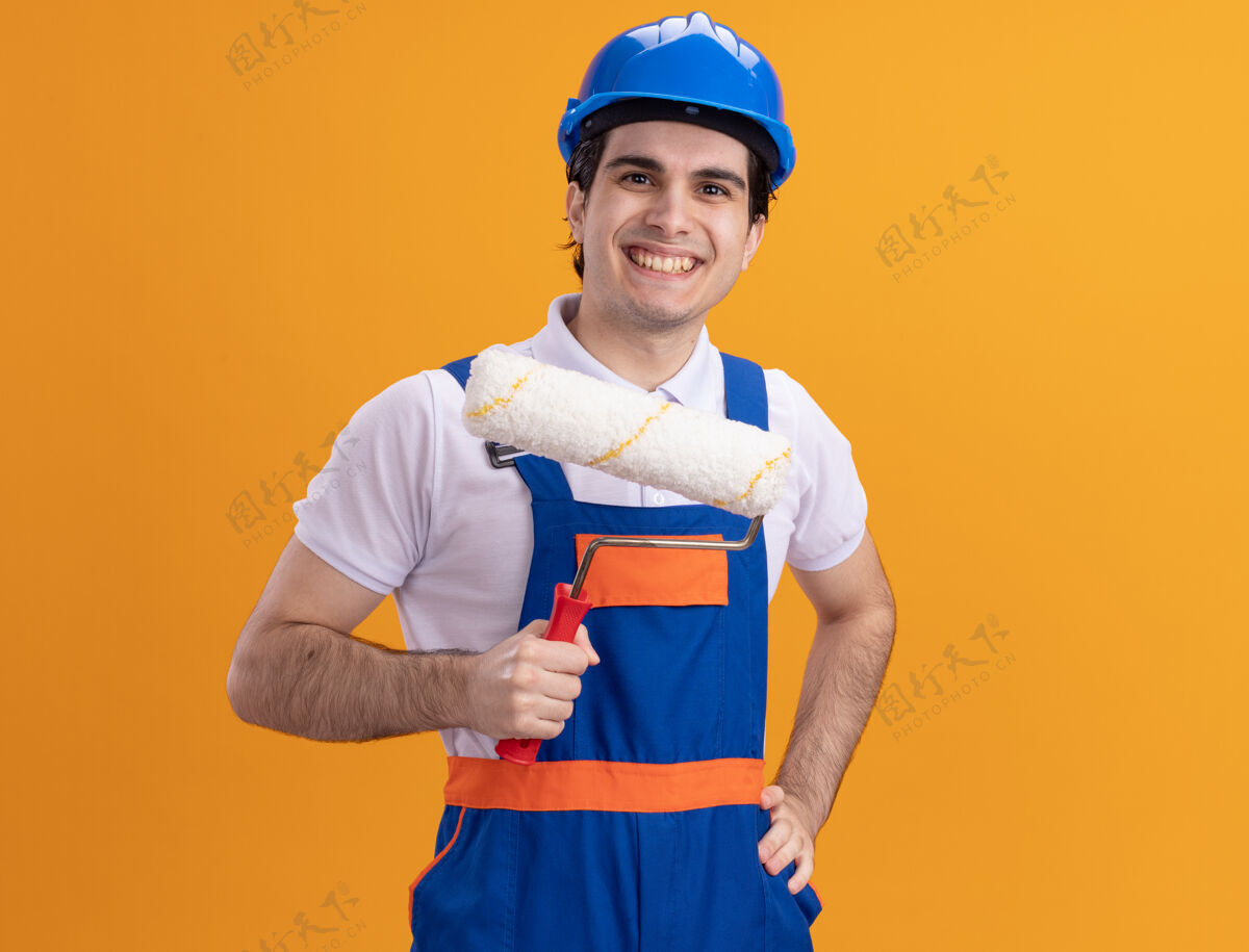 头盔年轻的建筑工人穿着建筑制服 戴着安全帽 拿着油漆辊 面带微笑地站在橙色的墙上 看着前面安全年轻人站着