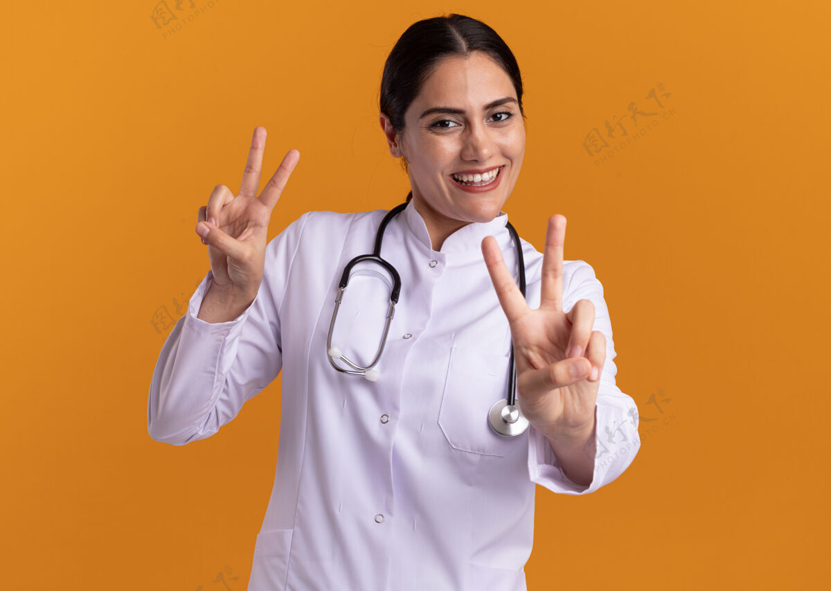 手势身穿医用外套的年轻女医生 脖子上戴着听诊器 面带微笑地看着前方 橙色的墙上立着v字标志脸围着医生