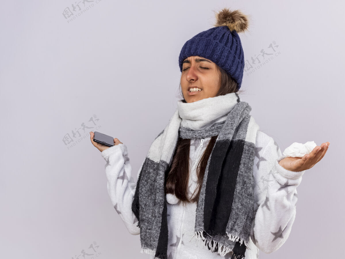 女孩关心的年轻病女孩闭着眼睛戴着冬天的帽子围巾拿着电话和餐巾 摊开双手隔离在白色背景上封闭围巾传播