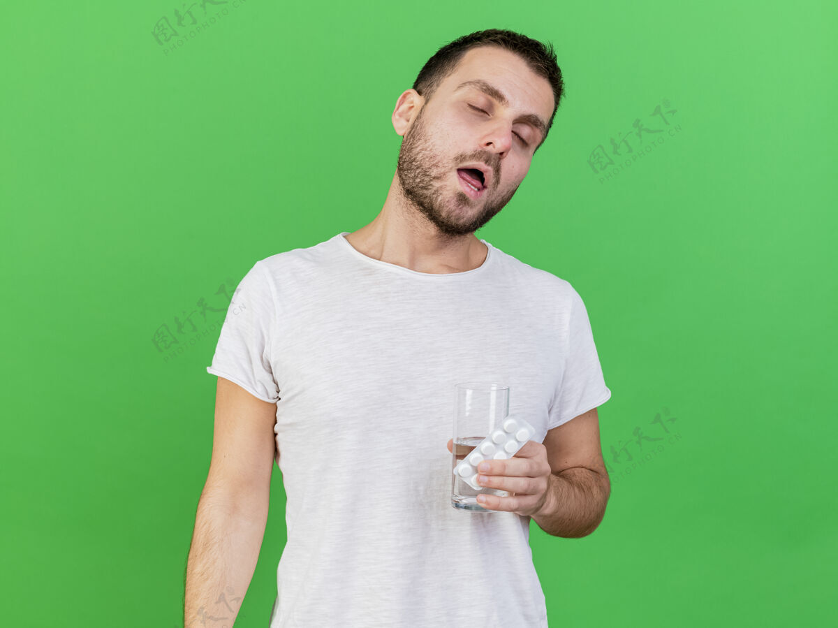 闭上年轻的病人闭着眼睛拿着药片 一杯水隔离在绿色的背景上生病年轻玻璃