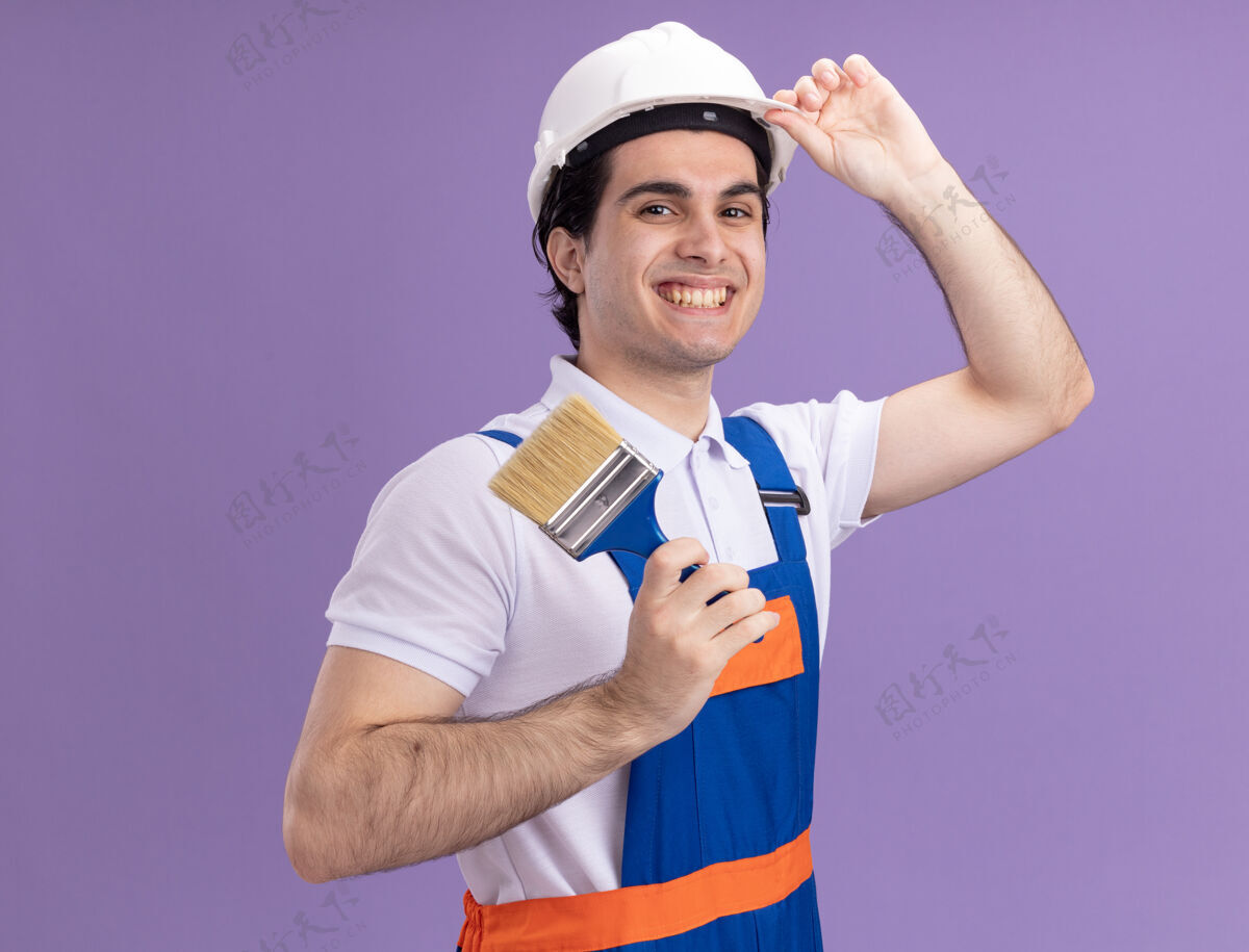 建筑快乐的年轻建筑工人穿着建筑制服 戴着安全帽 拿着油漆刷 微笑着自信地站在紫色的墙上 看着前方抱着刷子头盔