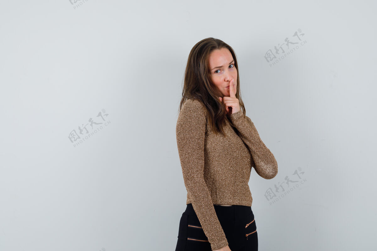 自然一个穿着金色毛衣和黑色裤子的年轻女子 用食指捂着嘴 表现出沉默的姿态 表情严肃年轻成人毛衣