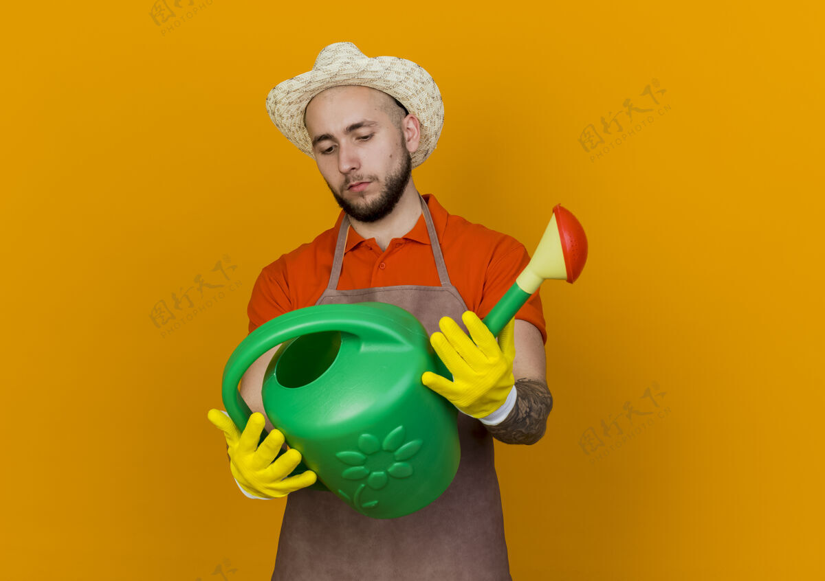 信心自信的男园丁戴着园艺帽拿着并看着浇水罐举行帽子橙色