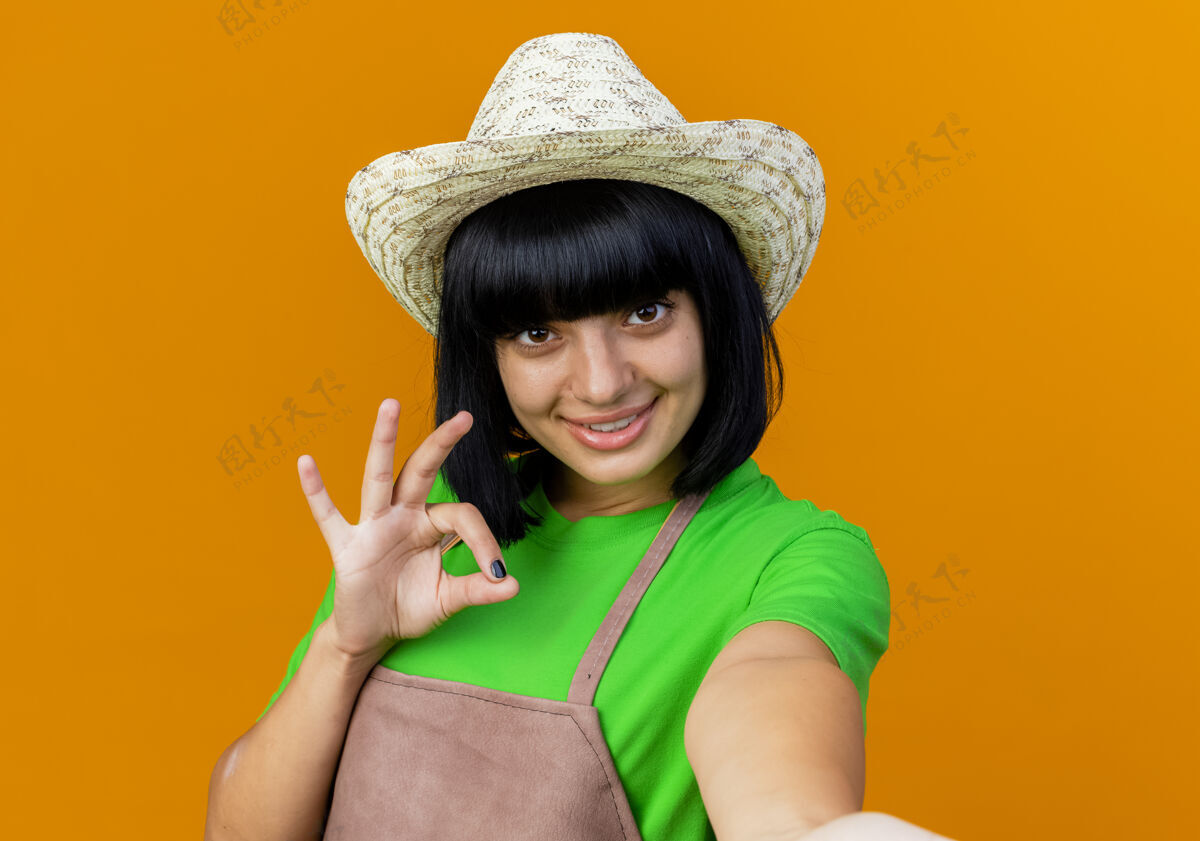 Ok微笑着的年轻女园丁身着制服 戴着园艺帽 手势ok 手势假装拿着相机假装花园复制
