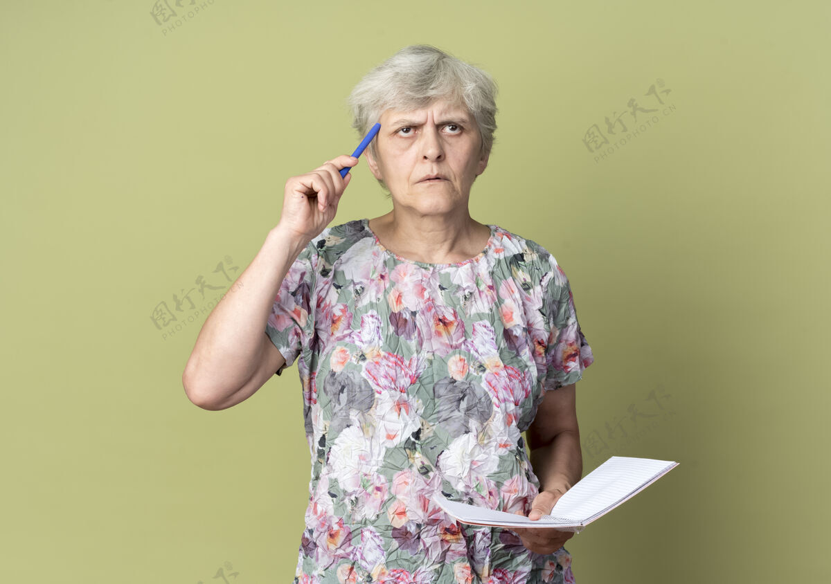 思想体贴的老妇人拿着笔记本 把笔放在橄榄绿墙上的圣殿上女人老人笔记本