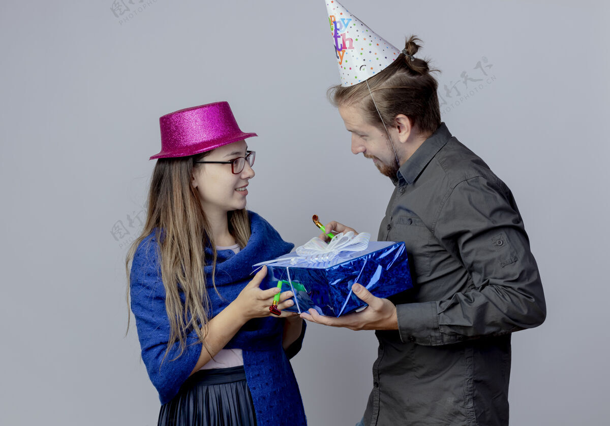 女人高兴的年轻夫妇互相看着对方拿着礼盒戴着粉色帽子戴着粉红色帽子的女孩拿着口哨和戴着生日帽的帅哥拿着口哨孤立在白色的墙上礼物帽子男人