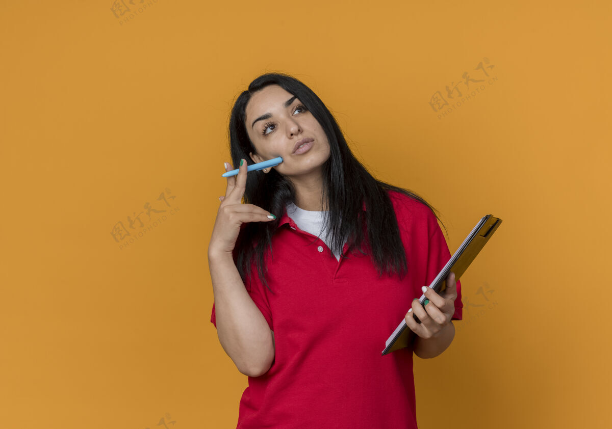 橙色体贴的年轻黑发白人女孩穿着红衬衫拿着笔和剪贴板孤立在橙色的墙上穿着剪贴簿钢笔