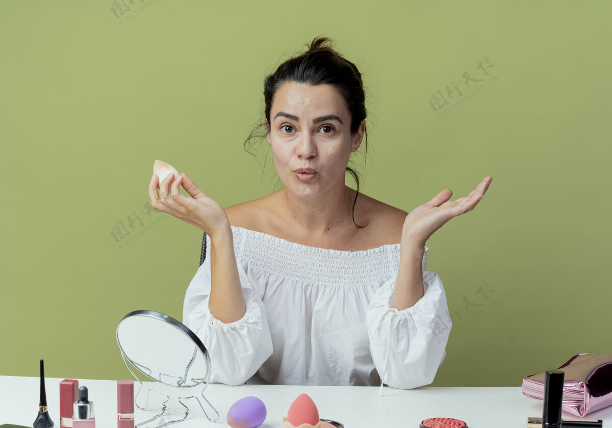 化妆惊艳的美女坐在桌旁用化妆工具用海绵打基础 看着绿墙隔开桌子海绵基础