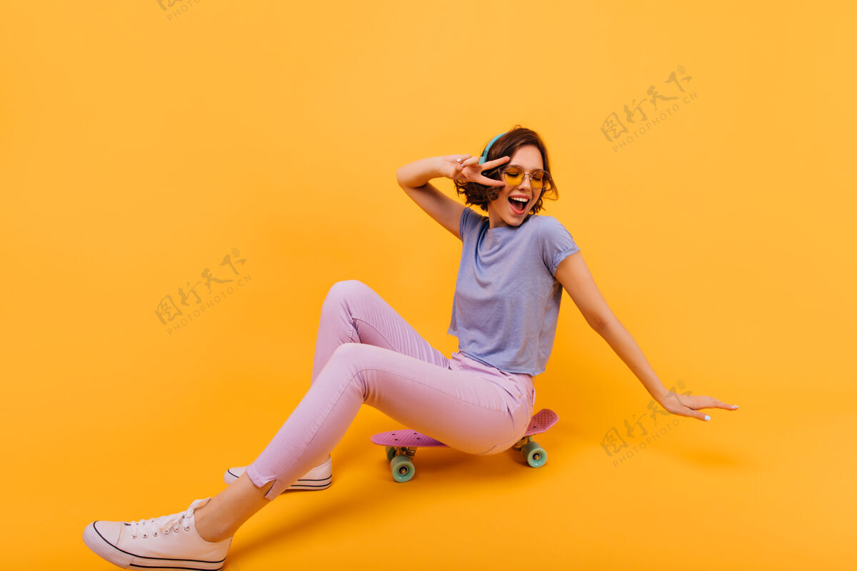 人温文尔雅的白人女孩穿着白色的鞋子坐在滑板上戴着耳机的笑容可掬的美女摆姿势乐趣玩热情