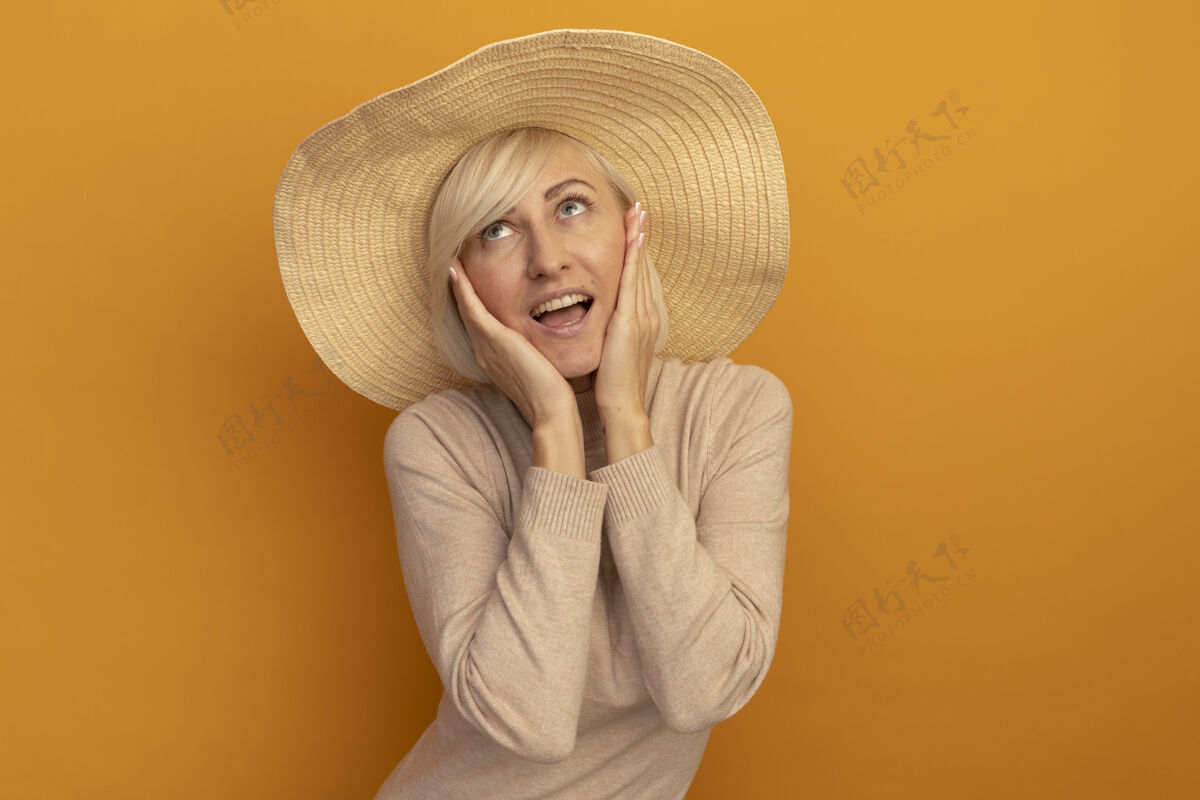 成人戴着沙滩帽的漂亮金发斯拉夫女人把手放在脸上 抬起头看着橙色手女人海滩