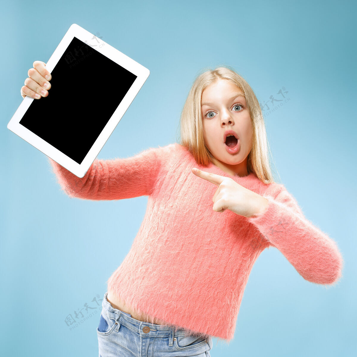 惊喜在蓝色画室背景上拿着平板电脑的有趣的小女孩她展示了一些东西并指着屏幕童年无线年轻
