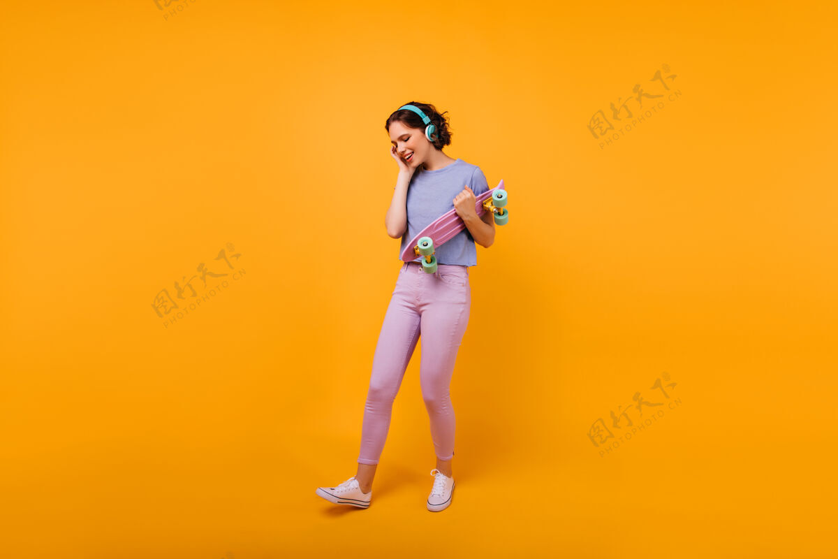 时尚穿着五颜六色休闲装的身材匀称的女孩戴着耳机听音乐可爱的短发女模特手拿着滑板长板活跃微笑