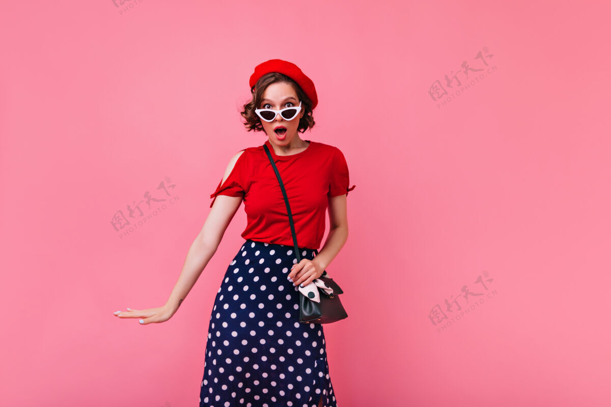 女性穿着红色贝雷帽的可爱女孩站在玫瑰色的墙上令人惊讶的法国女士波浪式发型的室内照片放松复古优雅