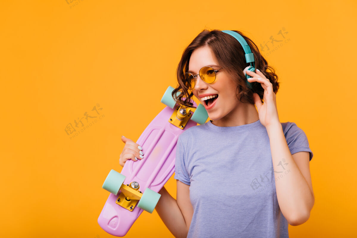 女性戴着黄色太阳镜的漂亮女孩戴着耳机听音乐穿着滑板唱着最喜欢的歌的时尚女模特的肖像酷放松无忧无虑