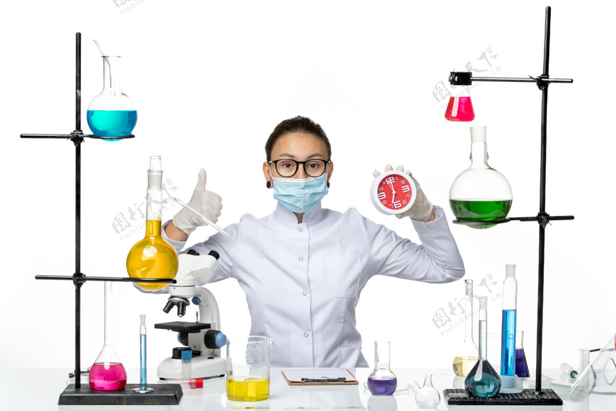 面具前视图穿着医疗服的女化学家 戴着面罩 在浅白色背景上拿着时钟病毒实验室化学covid-splash持有研究查看