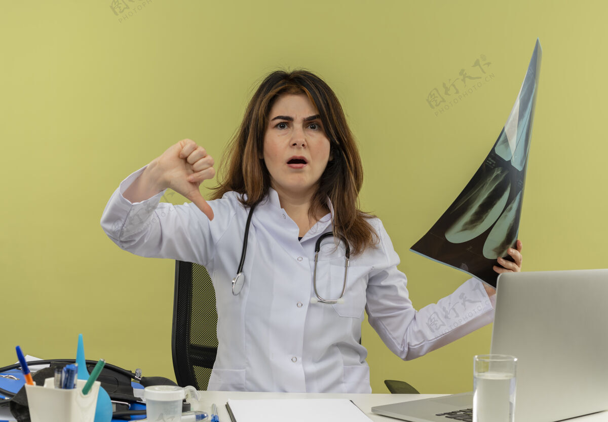 拇指中年女医生穿着医用长袍和听诊器坐在办公桌旁 手持医疗工具和手提电脑 手持x光片 显示拇指被隔离拍摄成人按住