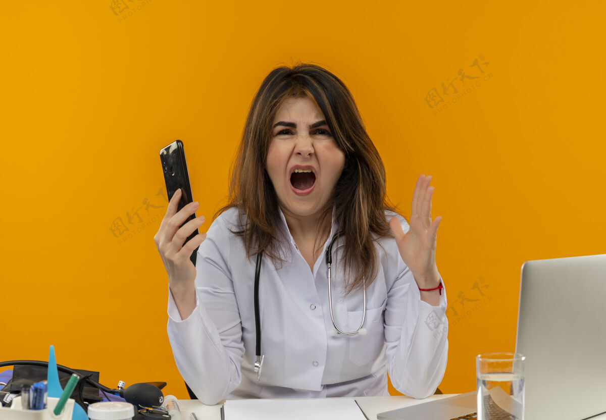 空气愤怒的中年女医生穿着医用长袍和听诊器坐在办公桌旁 手里拿着医疗工具 剪贴板和笔记本电脑 手里拿着手机 手与空气隔绝医生电话坐着