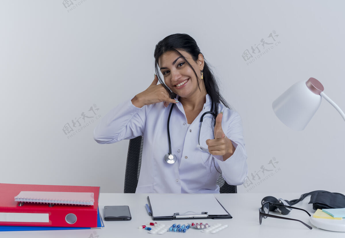 电话面带微笑的年轻女医生穿着医用长袍和听诊器坐在办公桌旁 手里拿着医疗工具 做着呼叫手势和手枪手势坐着桌子长袍