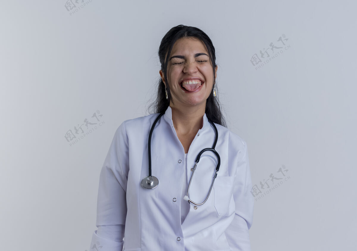 听诊器顽皮的年轻女医生穿着医用长袍和听诊器 闭着眼睛看舌头女人医生眼睛