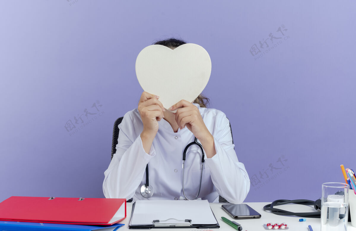 人年轻的女医生穿着医用长袍和听诊器坐在办公桌旁 手持医疗工具 躲在心形隔离的背后桌子听诊器穿