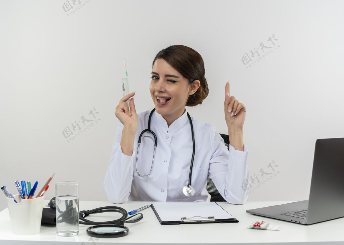 女士顽皮的年轻女医生穿着医用长袍和听诊器坐在办公桌旁 手里拿着医疗工具和笔记本电脑 手里拿着注射器 眨着眼睛 露出舌头 竖起手指女性抱着手指