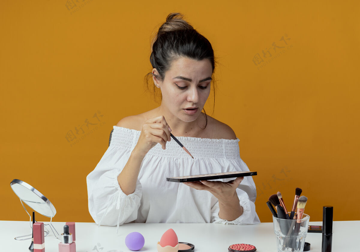 眼影自信漂亮的女孩坐在桌旁 用化妆工具拿着化妆刷 看着墙上的眼影调色板隔离桌子美丽化妆品
