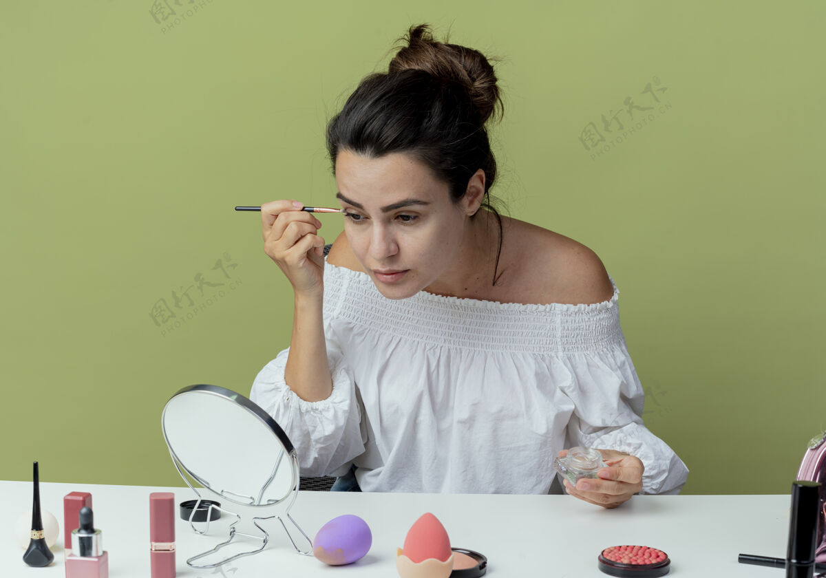 工具自信美丽的女孩坐在桌旁用化妆工具看着镜子 用眼影和化妆刷隔开绿色的墙美丽涂抹自信