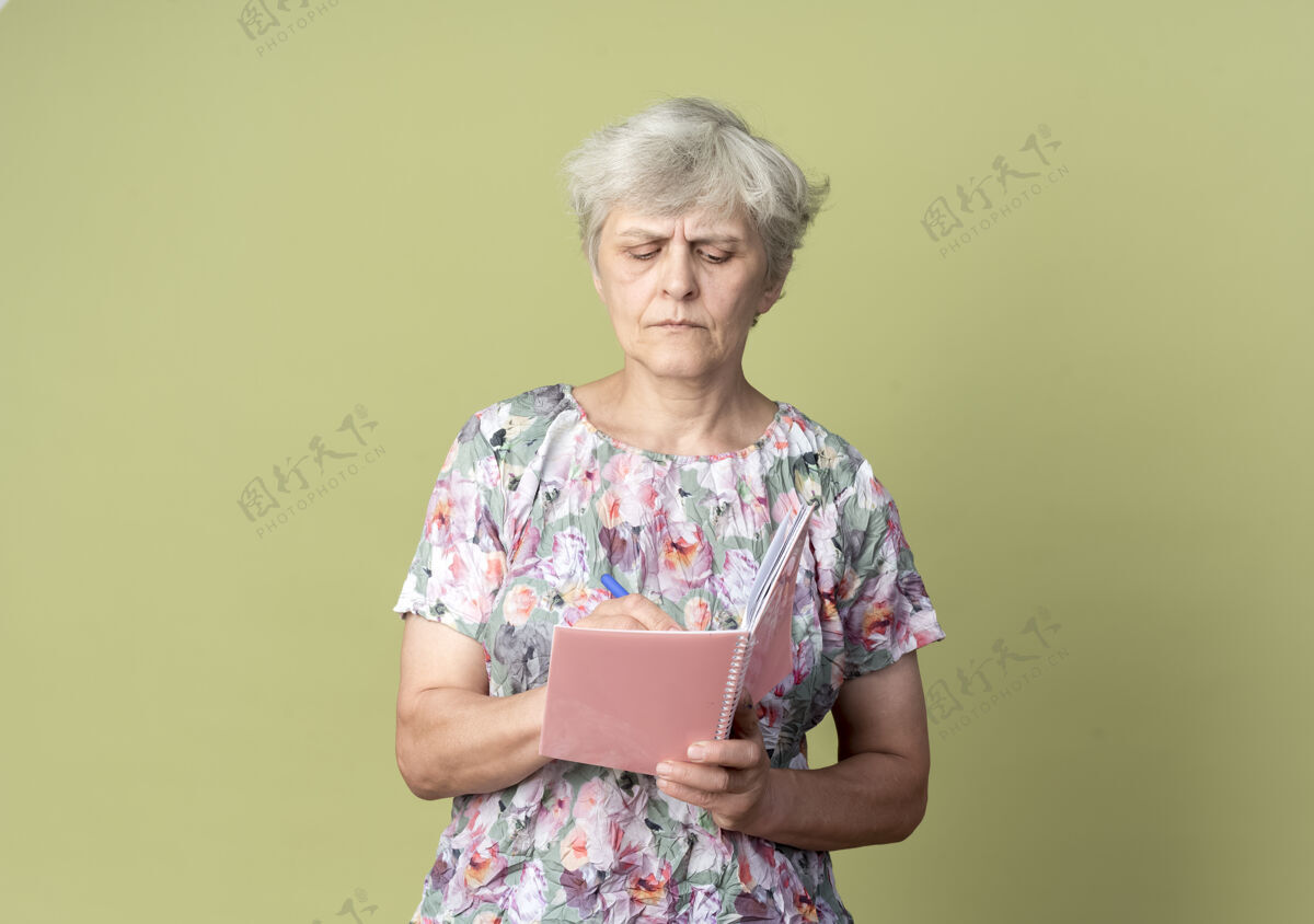 绿色自信的老妇人拿着笔记本和钢笔在橄榄绿的墙上写下一些孤立的东西信心女人钢笔