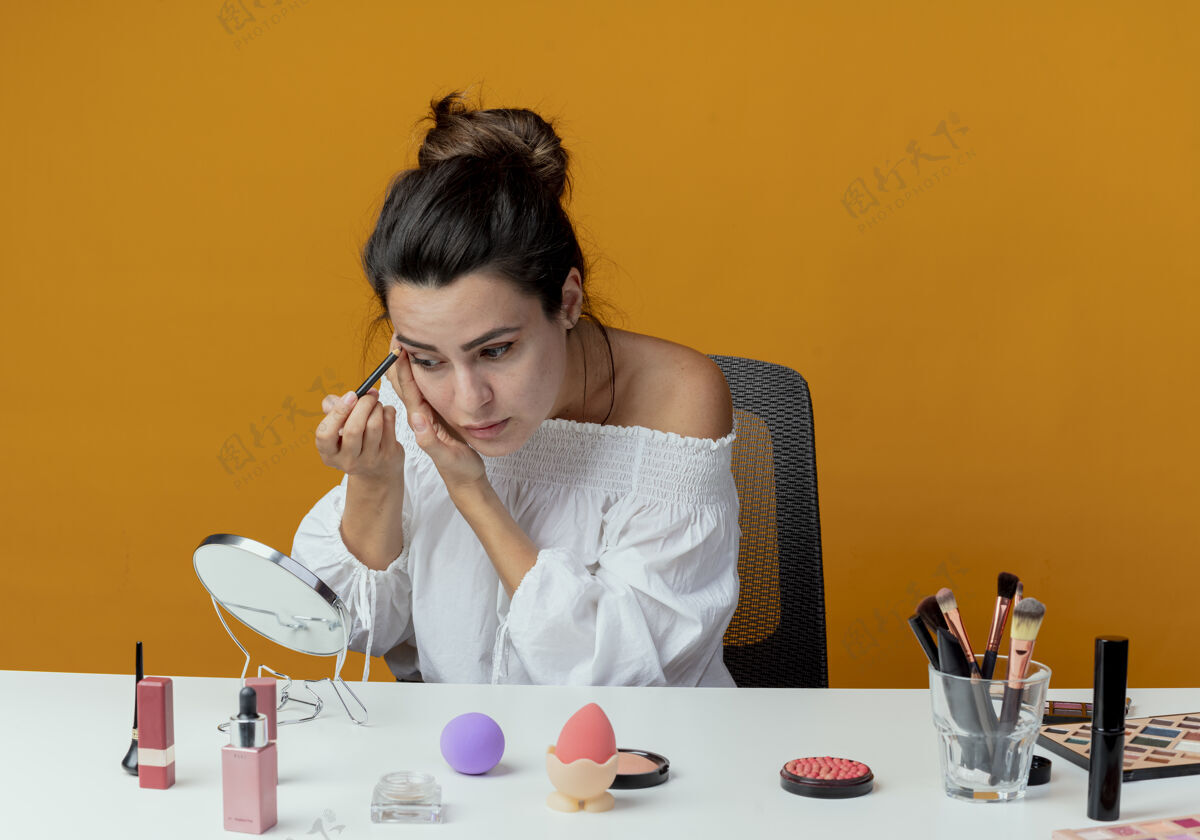橘子自信美丽的女孩坐在桌旁用化妆工具看着镜子 用眼影和化妆刷隔开橙色的墙壁女人镜子眼影