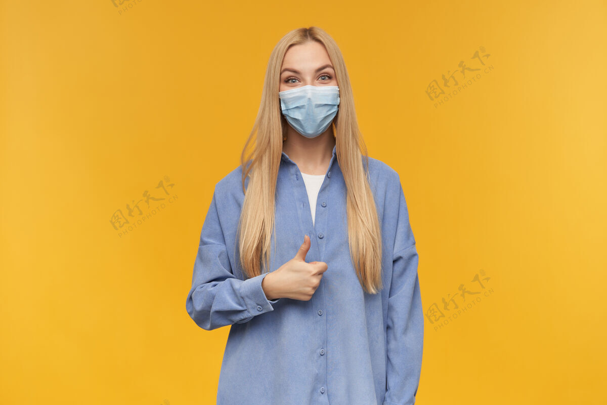 情绪漂亮的女人 漂亮的金发女孩竖起大拇指穿着蓝色衬衫和医用面罩橙色背景下隔离20多岁面具年轻