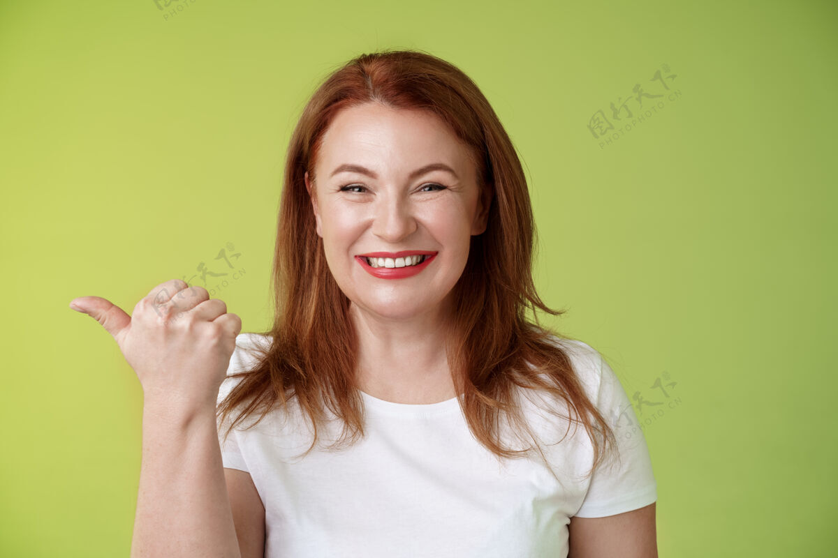 成熟欢迎光临本店开朗愉快友好迷人红发中年女企业家指着左手拇指站在绿色的墙上伟大化妆快乐