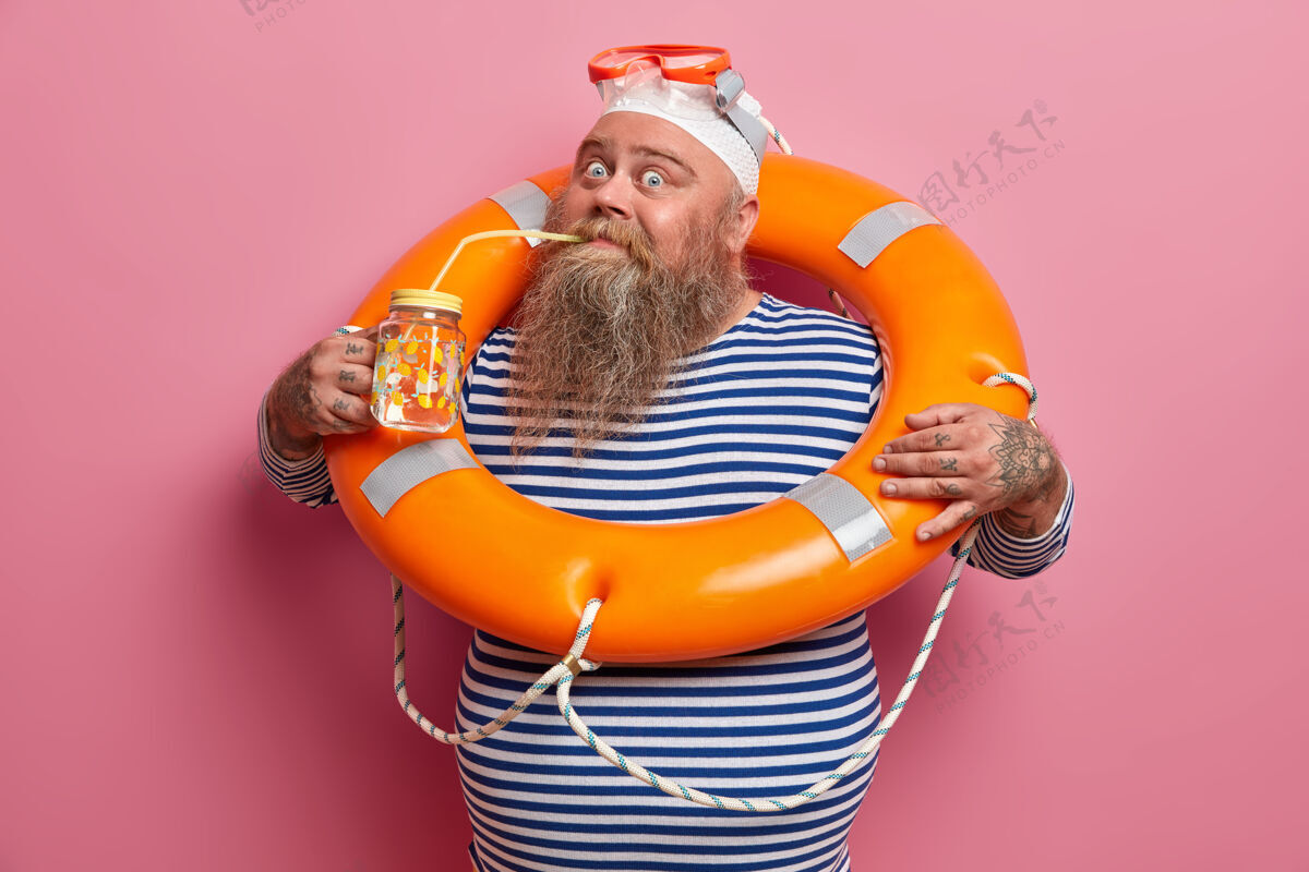 安全暑假和娱乐概念丰满的胡须成年男子在炎热的日子喝淡水 与救援橙色浮标摆姿势 戴泳帽和护目镜 隔离在粉红色的墙上救生员白种人穿着