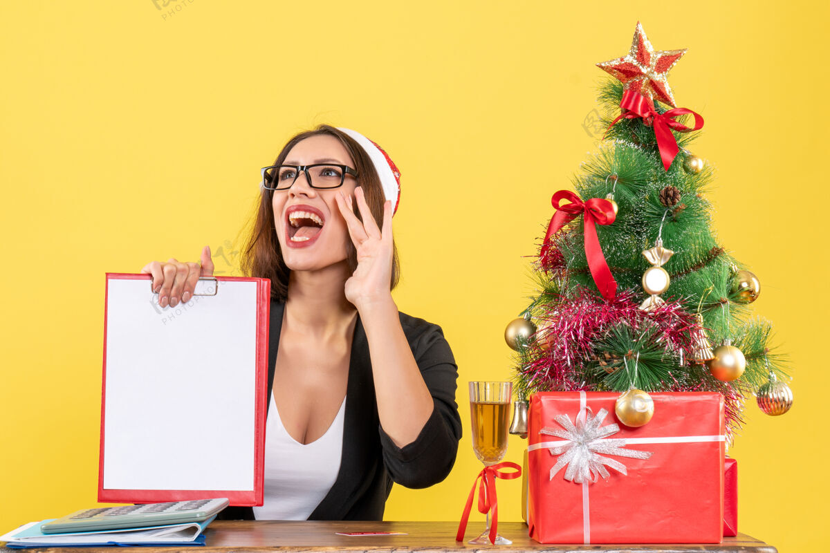 微笑微笑迷人的女士 穿着西装 戴着圣诞老人的帽子 戴着眼镜 展示着一份文件 上面写着在办公室里打电话给某人的黄色隔离带头发黄色帽子