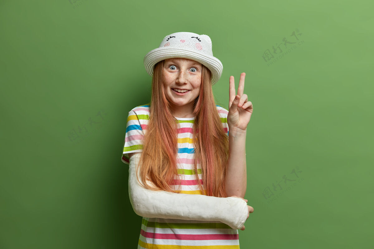 精力充沛开朗微笑的女孩 带着喜悦的表情 做出胜利或和平的手势 戴着帽子和条纹t恤 玩得很开心 手臂在石膏上断了 孤立在绿色的墙上孩子们 肢体语言少年积极快乐