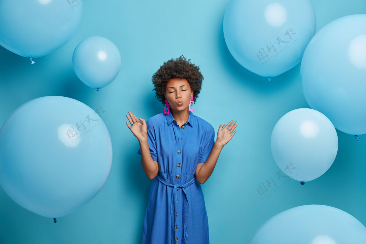 冷时尚迷人的女人举手 嘴唇圆润 心情愉快 拿着气球对着蓝色的墙壁摆姿势 在装饰好的房间里拍照 穿着漂亮的长裙明亮手掌衣服