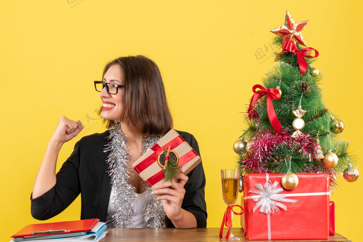 人一位身着西装 戴着眼镜 面带微笑的商务女士自豪地拿着礼物 坐在一张桌子旁 桌子上放着一棵圣诞树生活树套装