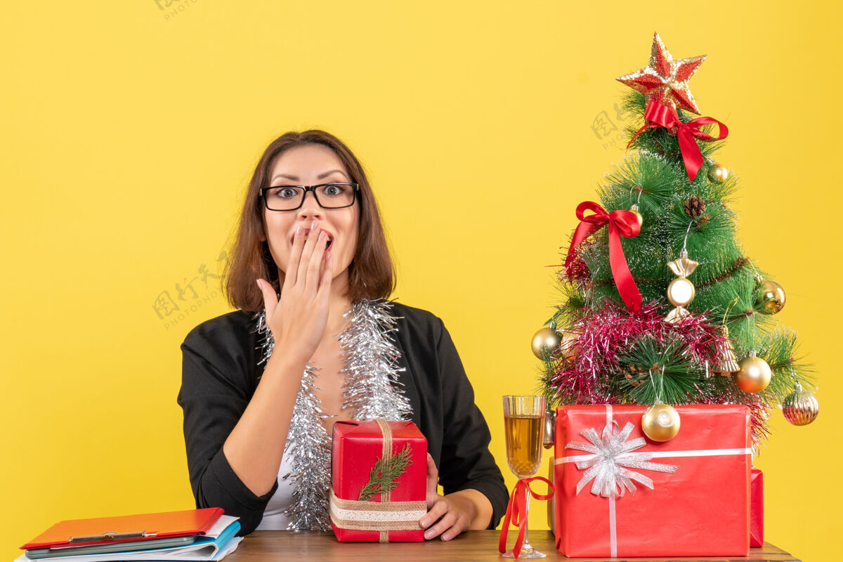 女士一位穿着西装 戴着眼镜 拿着礼物 坐在一张桌子旁 桌上放着圣诞树 情绪激动的商务女士在办公室里大吃一惊树套装美丽